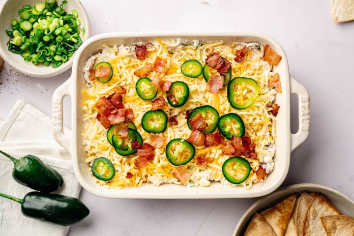 A rectangular baking dish filled with a cheesy dip, topped with sliced jalapeños and crispy bacon. Nearby are a bowl of chopped green onions, fresh jalapeños, and a bowl of tortilla chips.